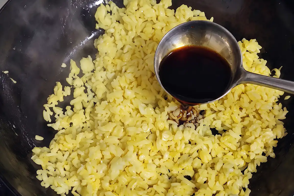 Drizzling light soy sauce from a ladle onto egg-coated fried rice with ham and shrimp