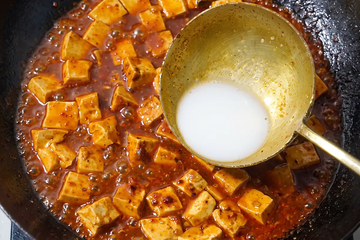 Adding white cornstarch slurry from a ladle to thicken the simmering Mapo Tofu sauce