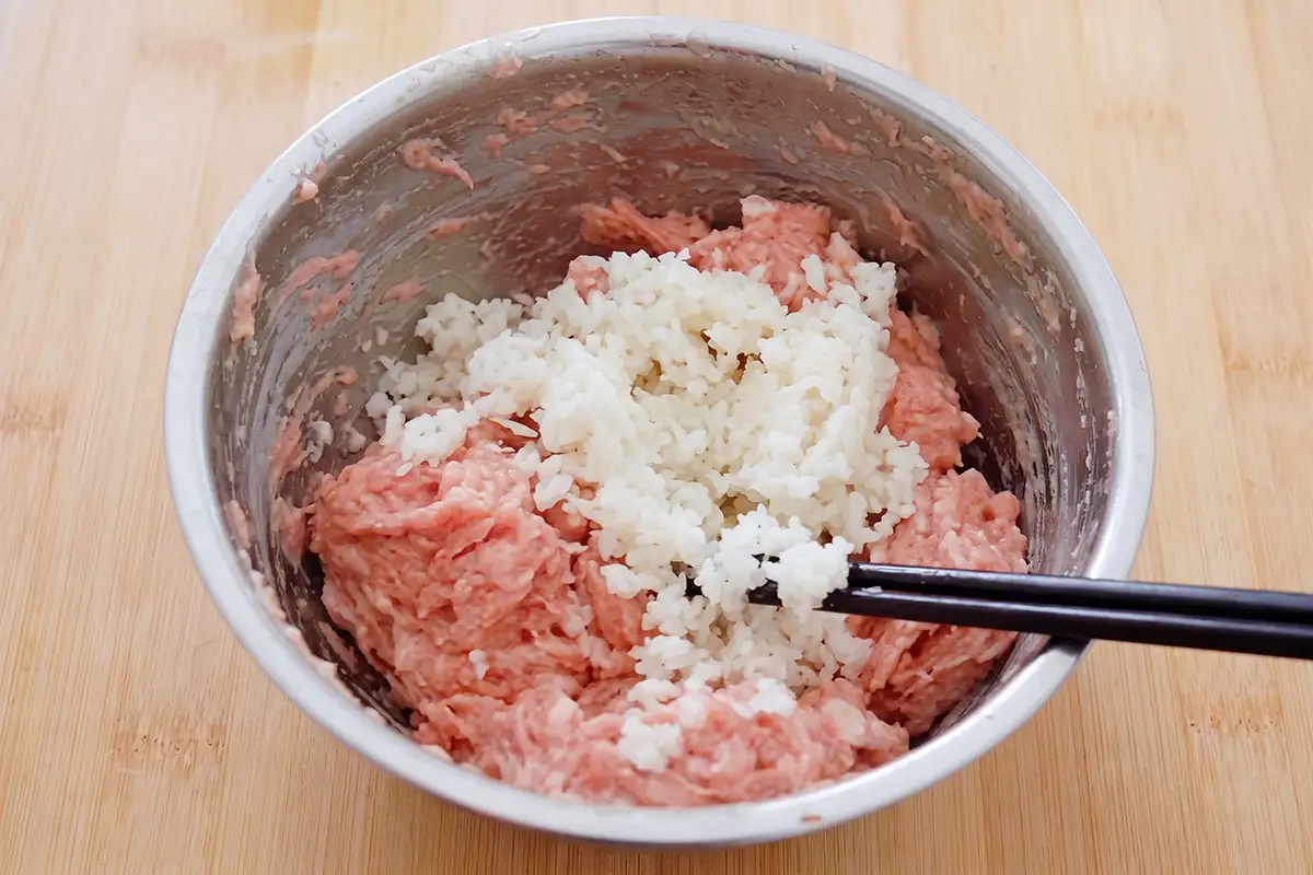 Finely diced water chestnuts being folded into hand-chopped pork with chopsticks in a stainless steel bowl