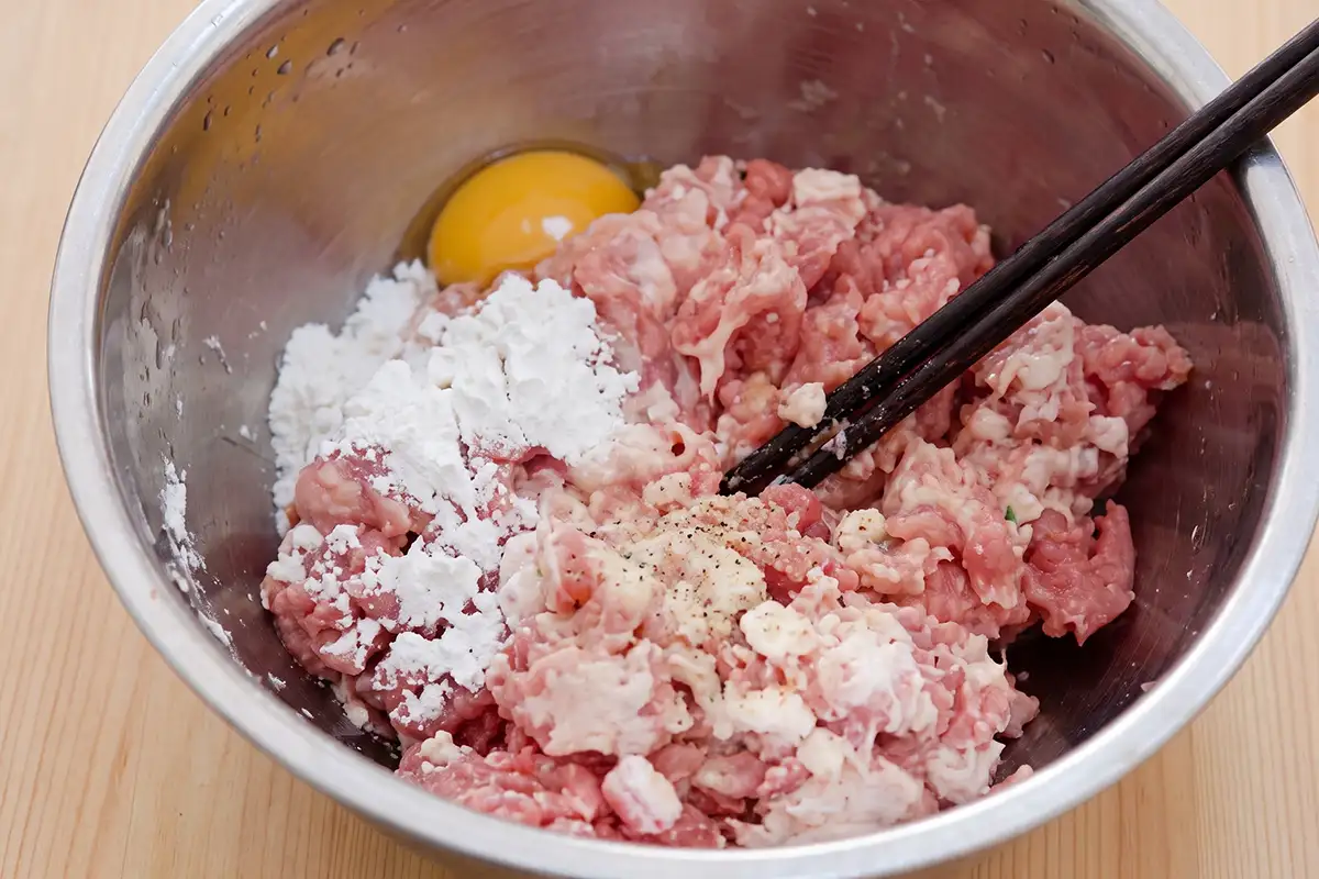 Ground pork with egg white, potato starch, and white pepper ready to be mixed in a stainless steel bowl