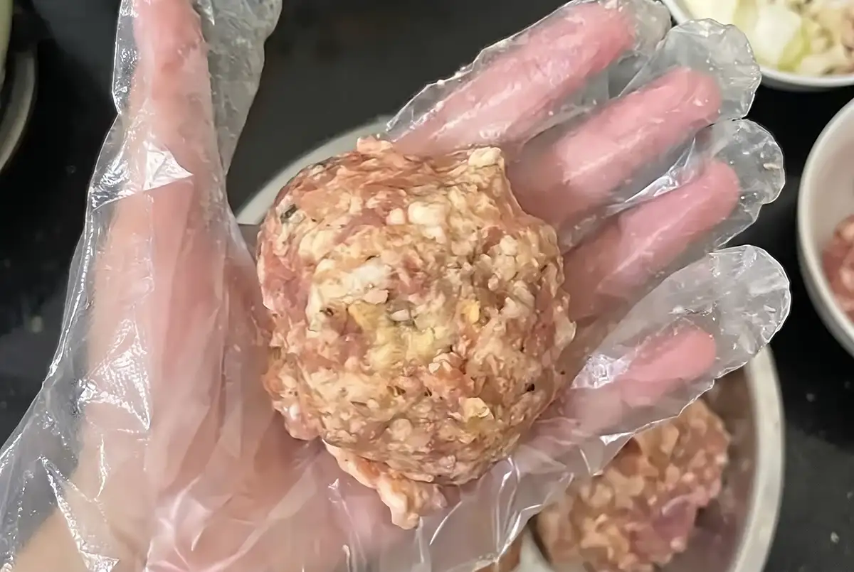 A large raw lion's head meatball gently shaped in a gloved hand, showing coarse, loose texture
